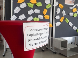 Ein Foto eines Schildes mit der Aufschrift: Schnapp dir einen Papierflieger und pinne deine Idee an die Zukunftswand! Dahinter ist eine Pinwand mit vielen angehefteten Papierfliegern zu sehen.